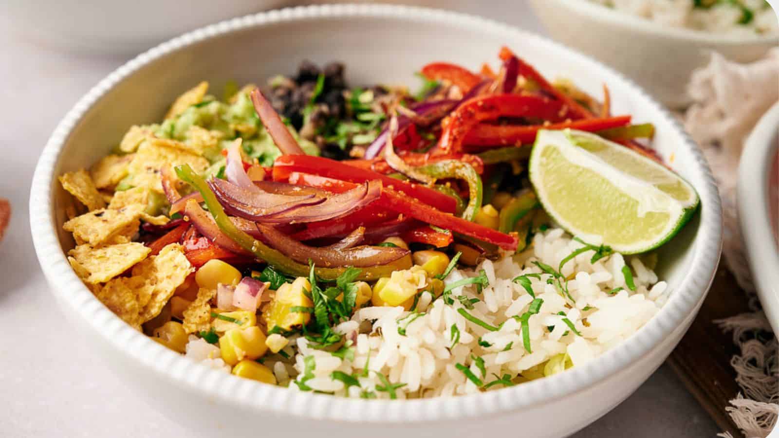 A Copycat Chipotle burrito bowl, filled with rice, fajita veggies, corn salsa, tortilla chips. and topped with a lime wedge.