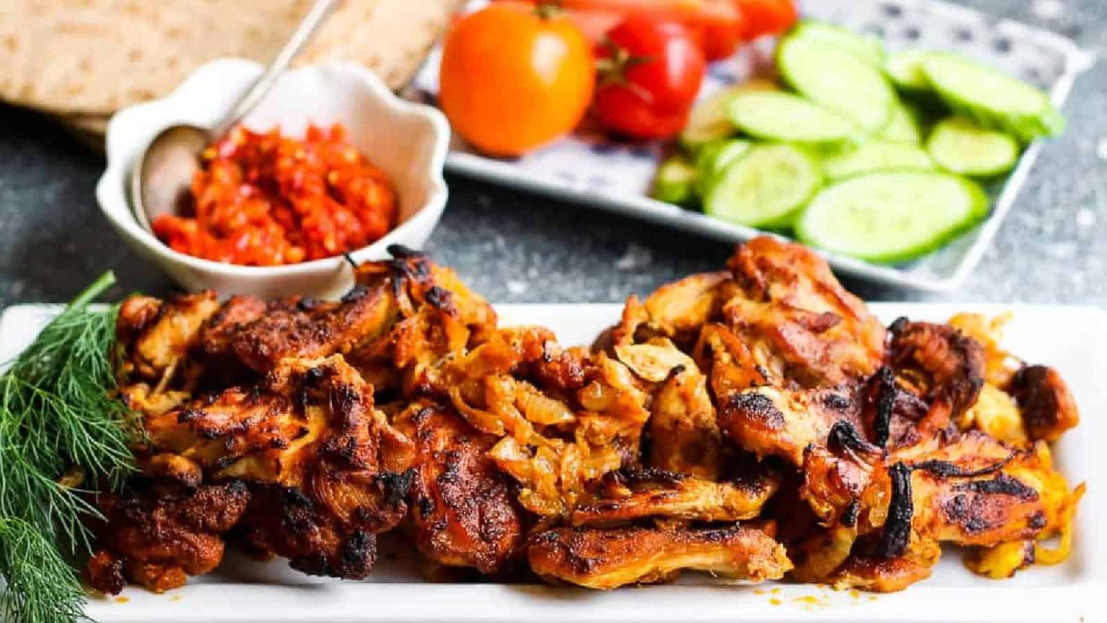 low angle shot of chicken shawarma on a white platter with harissa paste and tomatoes and cucumbers in the background.