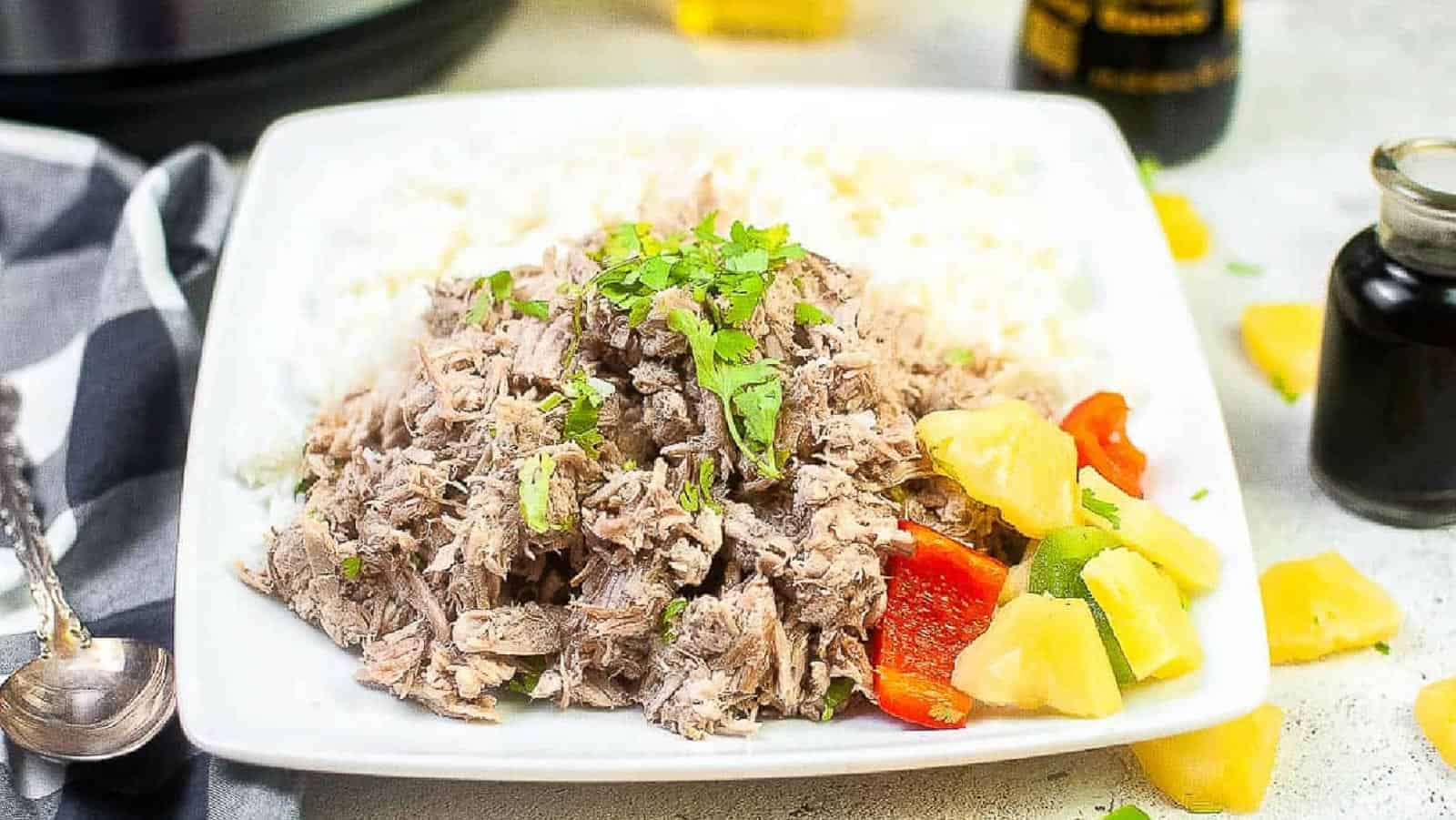 Low angle shot of kalua pork on a white plate with pineapple and pepper chunks.