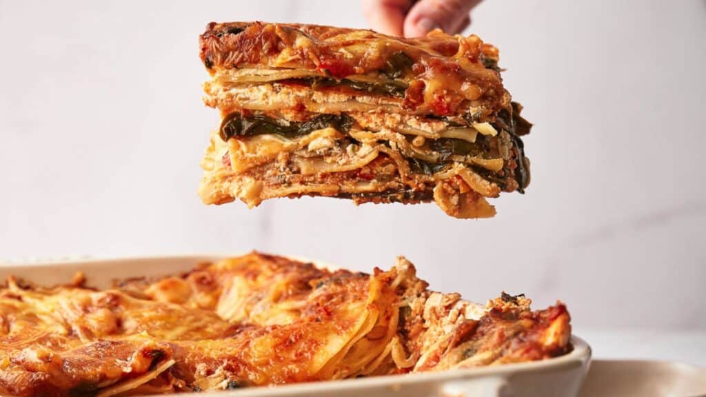 A person holding a slice of lasagna in a dish.