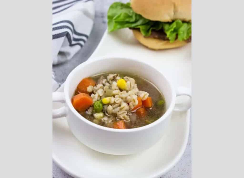 A bowl of rice and vegetable soup with a burger on a plate.