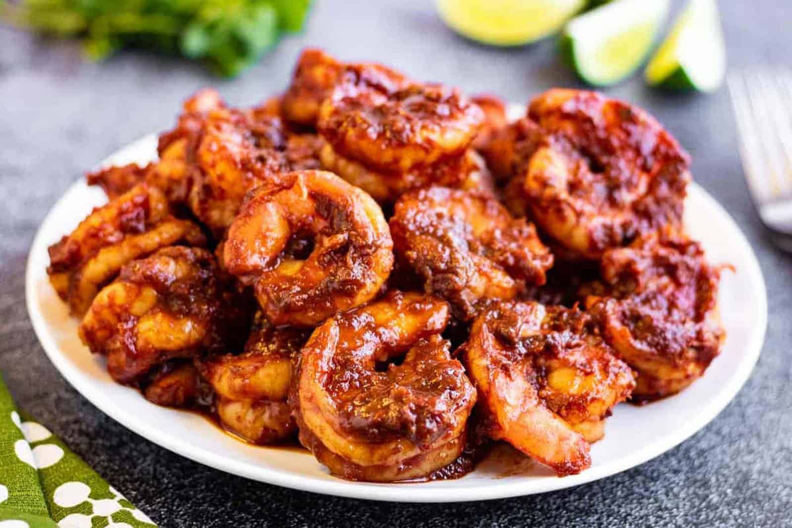 Shrimp in red chile sauce on a white plate with lime wedges and a fork in the background.