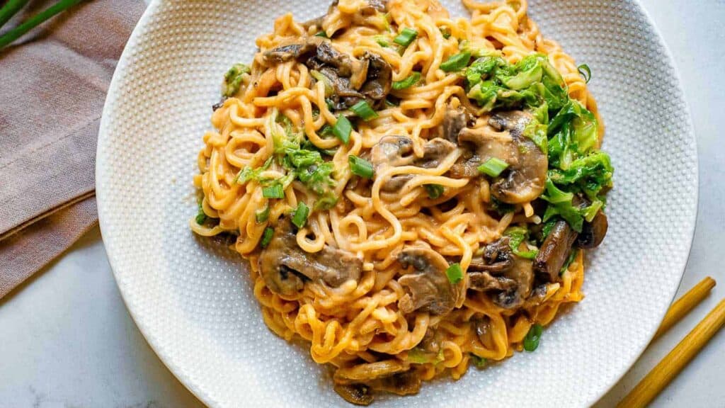 Low angle shot of gochujang noodles with greens, mushrooms, and cheese on a grey and white plate.