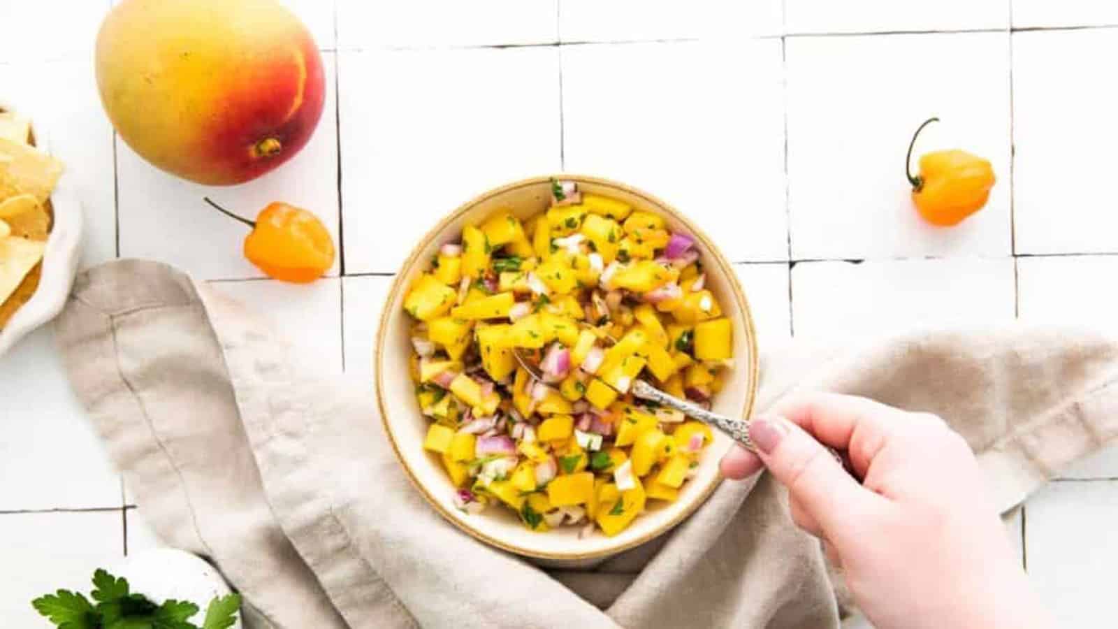 Overhead shot of a bowl of mango habanero salsa with a hand lifting a spoonful of the salsa.