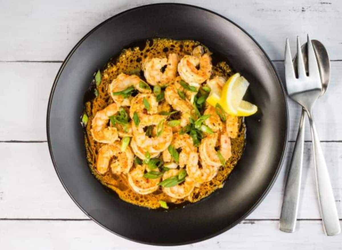 A plate with New Orleans BBQ Shrimp and lemon wedges, with serving utensils to the side.