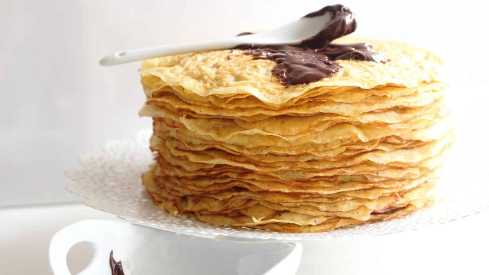 Crepes layered on top of each other on a cake stand with nutella on top. 