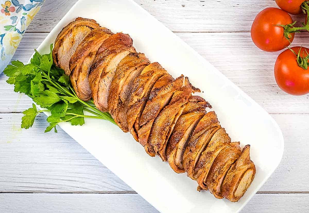 Sliced Smoked Bacon Wrapped Whole Pork Tenderloin on a white platter.