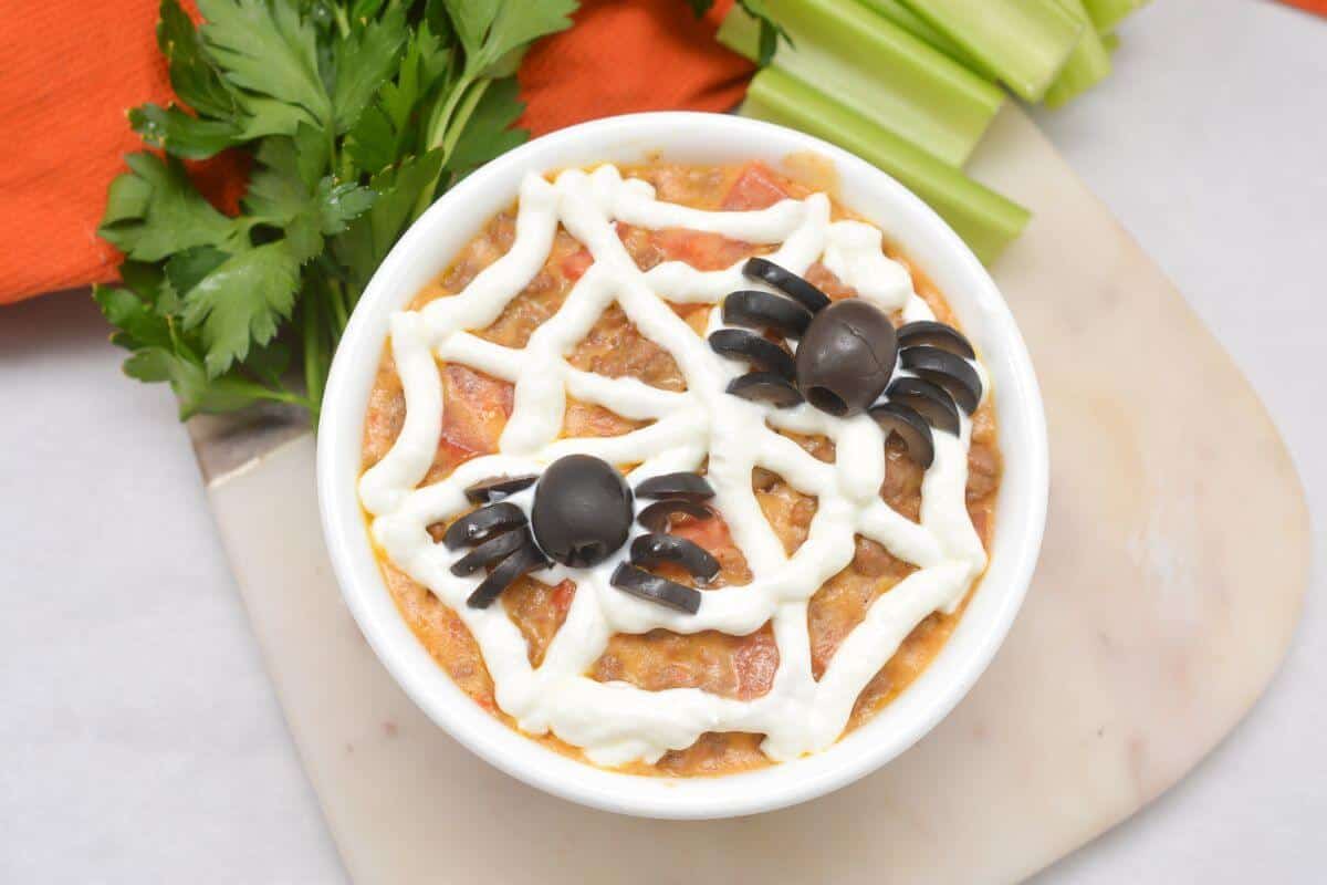 A bowl of beef queso dip with a spider web on top served with celery sticks.