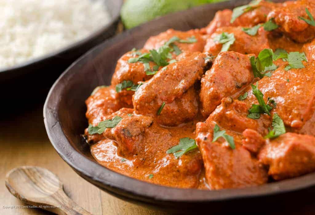 A bowl of chicken curry with rice and lime on a wooden table.