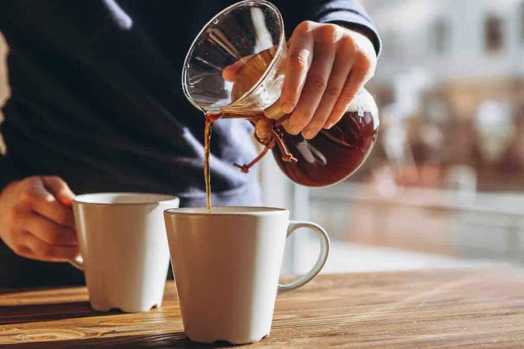 A person pouring coffee into two mugs on a wooden table.