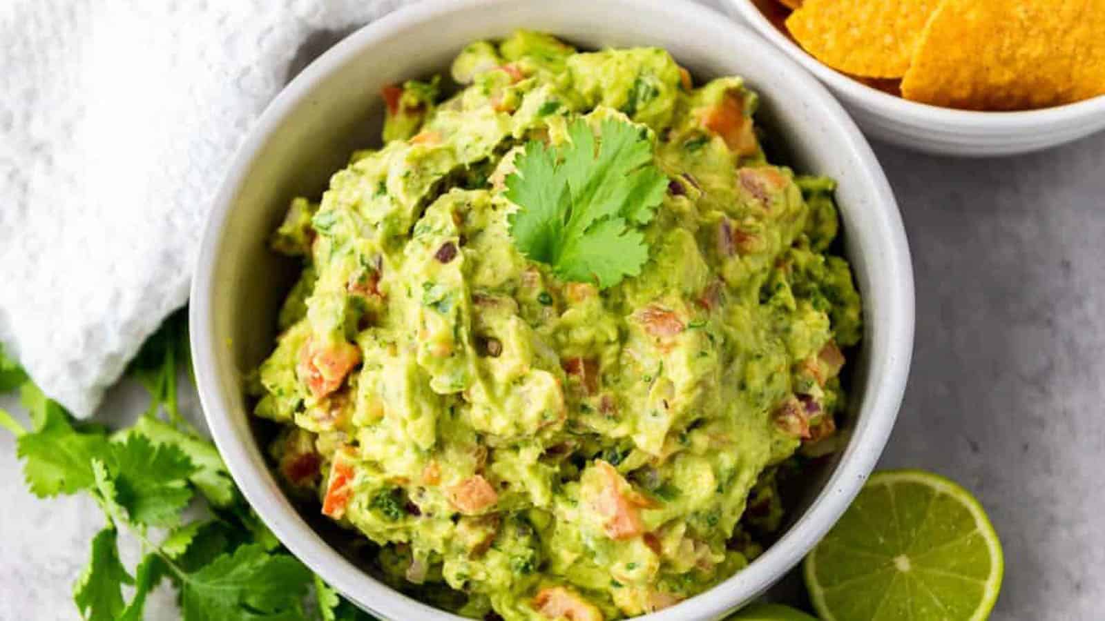 Guacamole in a white bowl with limes, cilantro, and tortilla chips on the side.