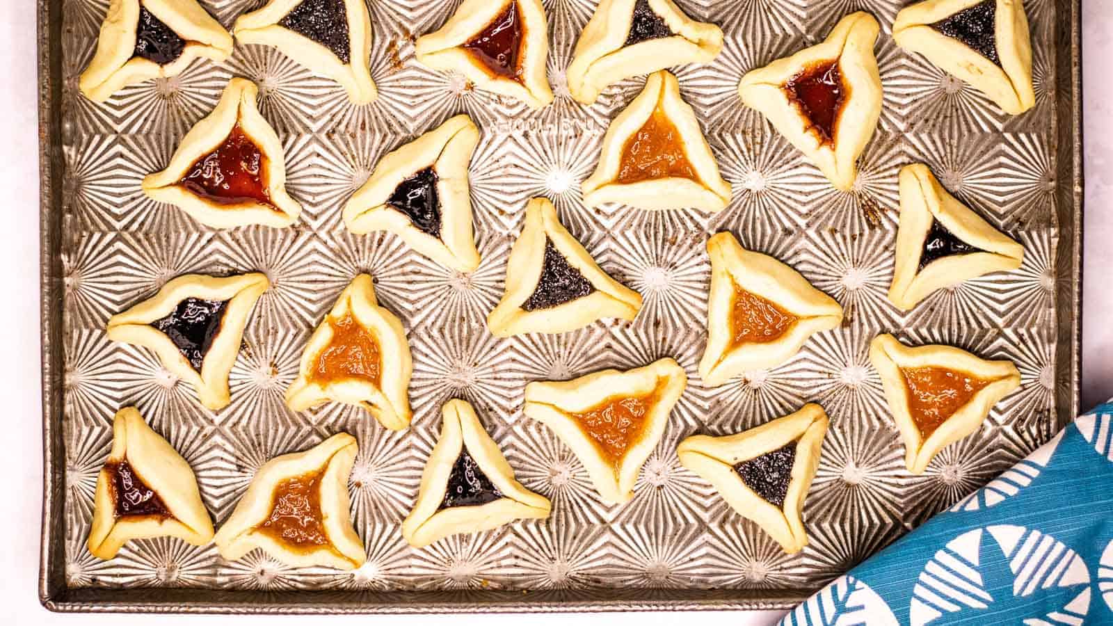 Hamentashen cookies piled on a white plate with more cookies on a rack in the background. There is jar of jam with a spoon in it in the background too.