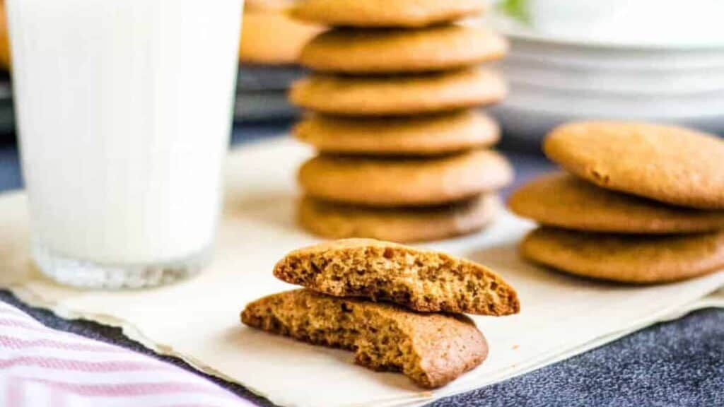 Low-angled shot of a honey cookie broken in half with stacks of honey cookies and a glass of milk in the background.