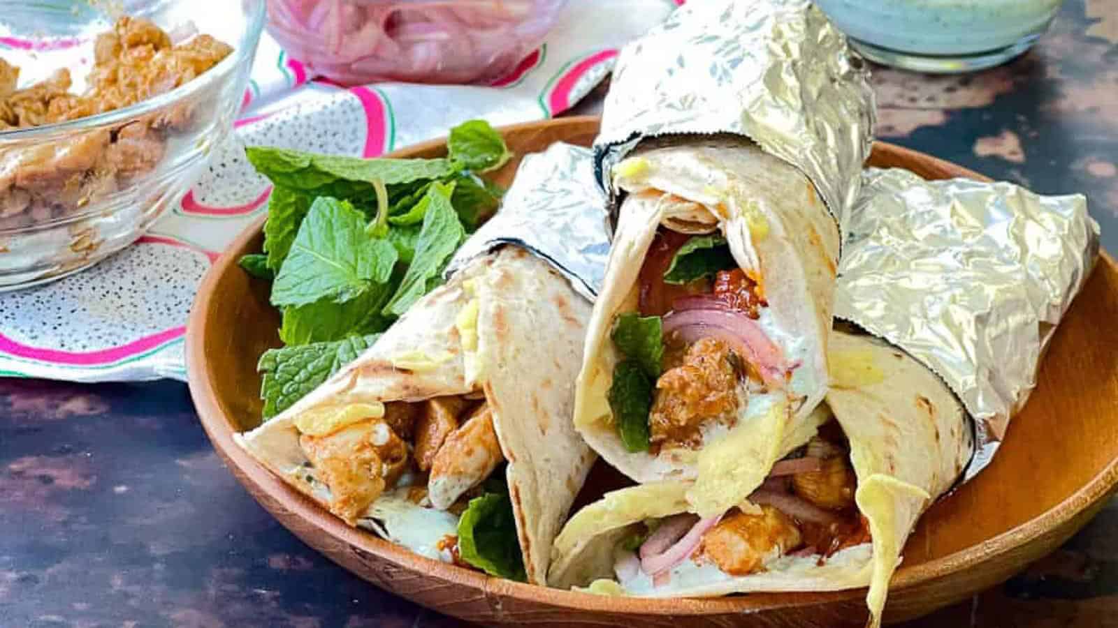 Low angle shot of 3 chicken kathi rolls wrappedin foil on a wooden plate with fresh mint leaves.