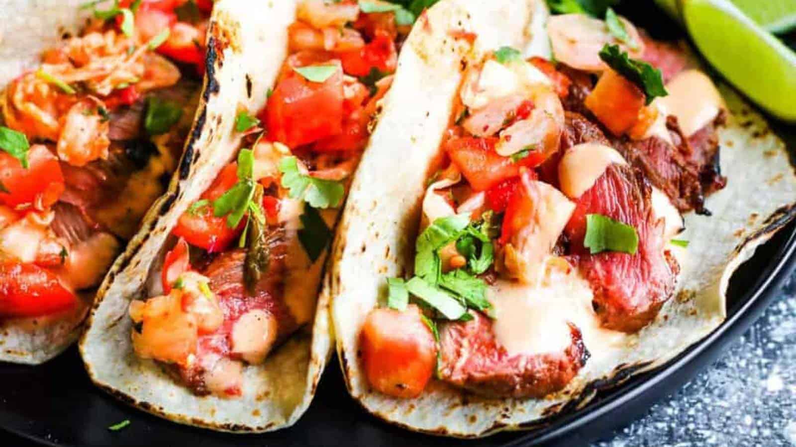 Horizontal shot of 3 korean tacos: corn tortillas filled with sliced steak, kimchi and pico de gallo salsa, and chopped cilantro. In the background are lime wedges, sliced red cabbage, and cilantro leaves.