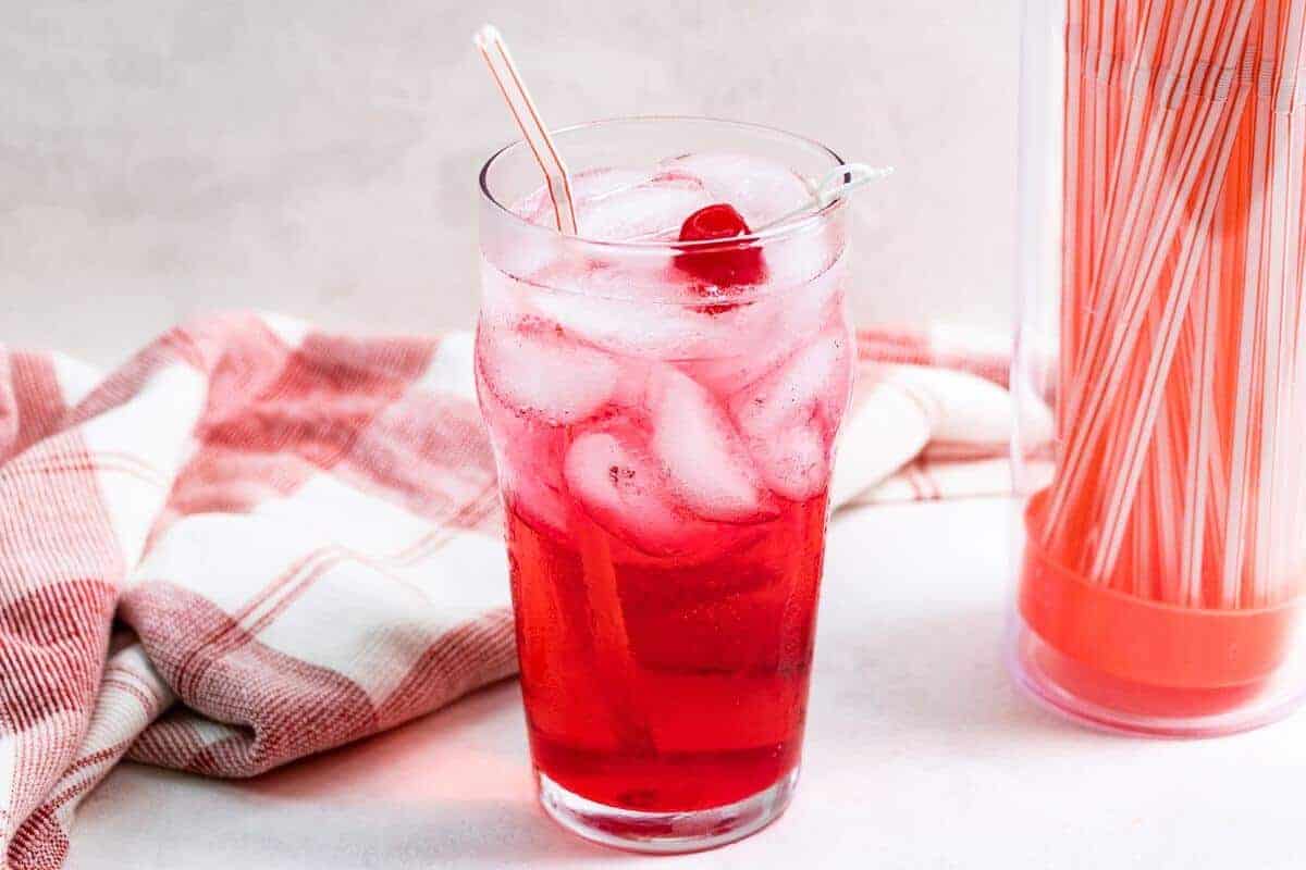 Shirley temple drink in tall glass with cherry garnish and straws container.