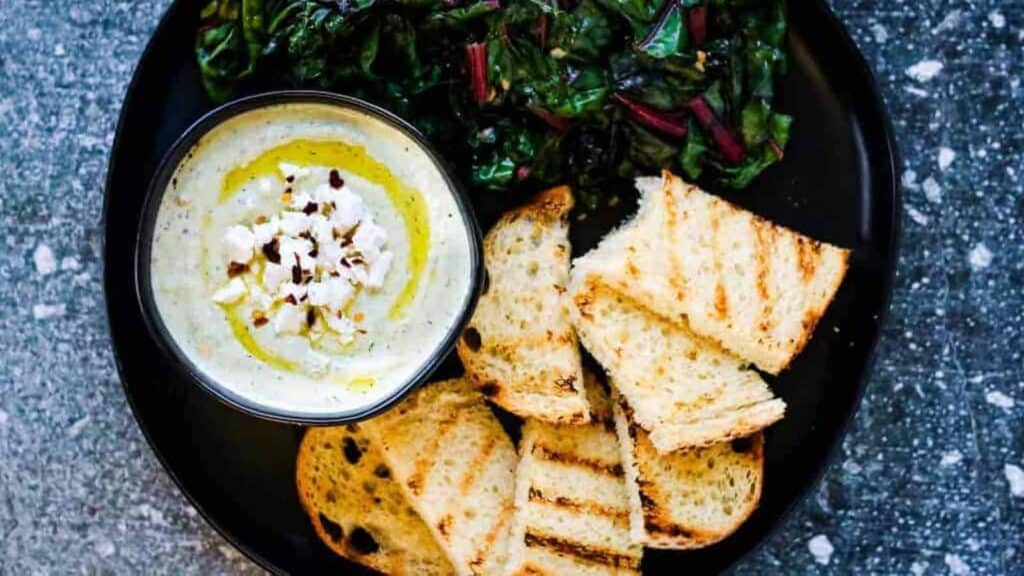 Overhead shot of a black platter with a bowl of whipped feta spread, sauteed chard, and grilled sourdough bread.