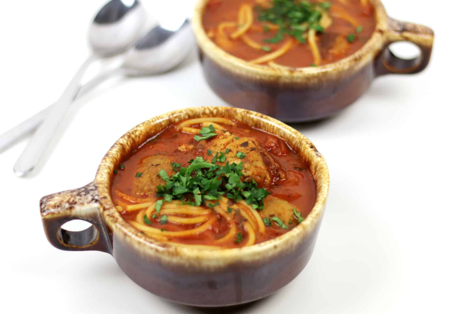 Two silver spoons and two bowls of spaghetti and meatballs soup in brown ceramic bowls.