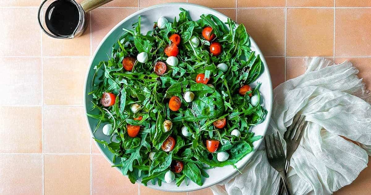 Arugula caprese salad on a white plate next to two forks, a white linen, and a small glass pitcher of balsamic glaze.