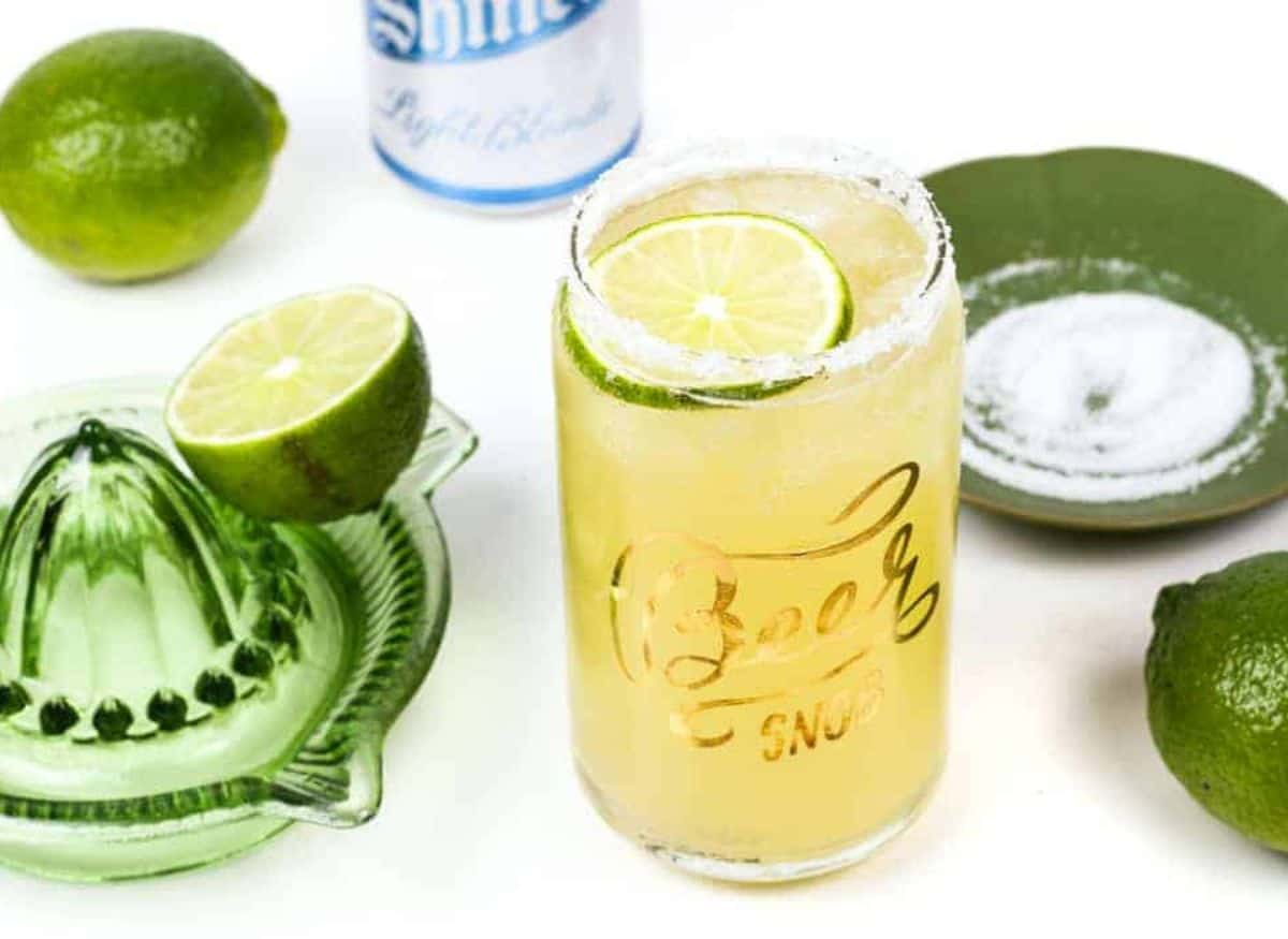 A beerita with a salt rim in a glass shaped like a beer can next to limes and a green glass juicer.