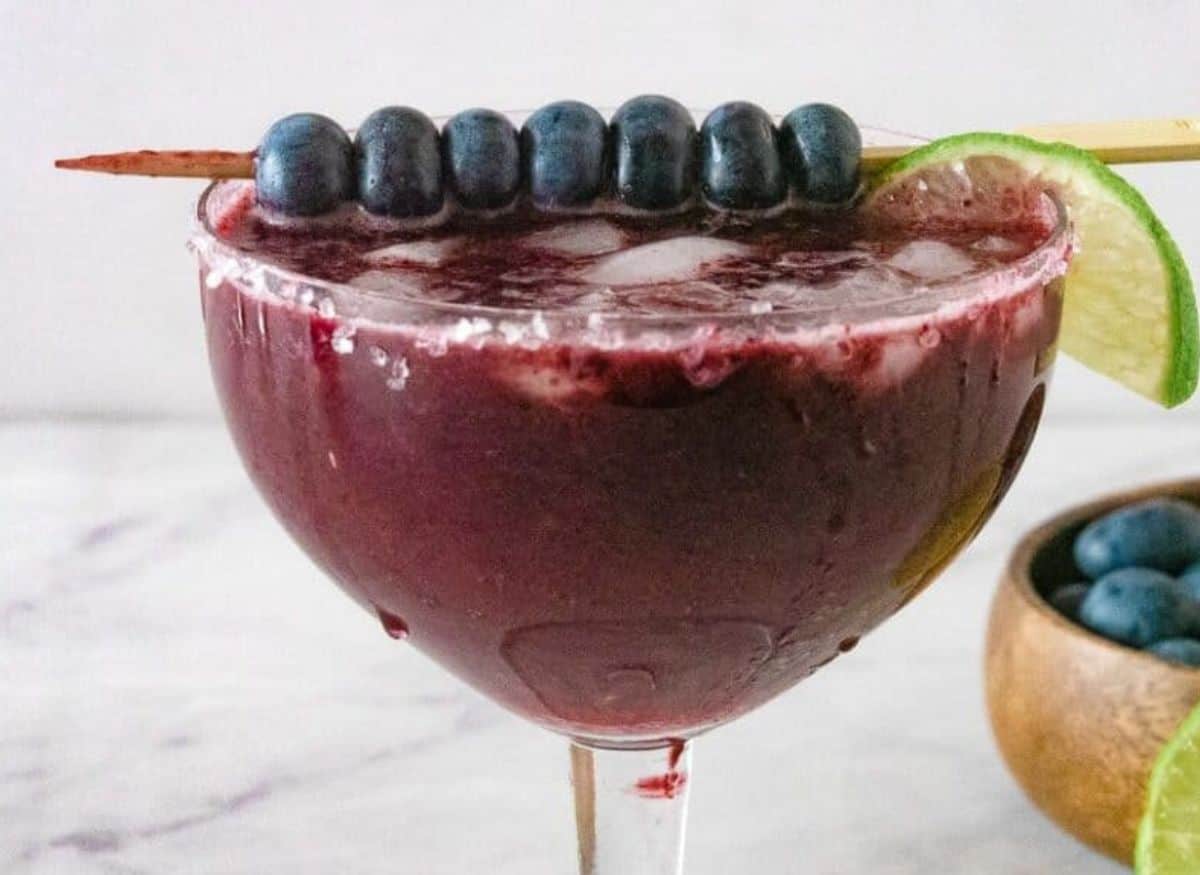 Margarita glass with blueberry margarita topped with lime and a skewer of blueberries.