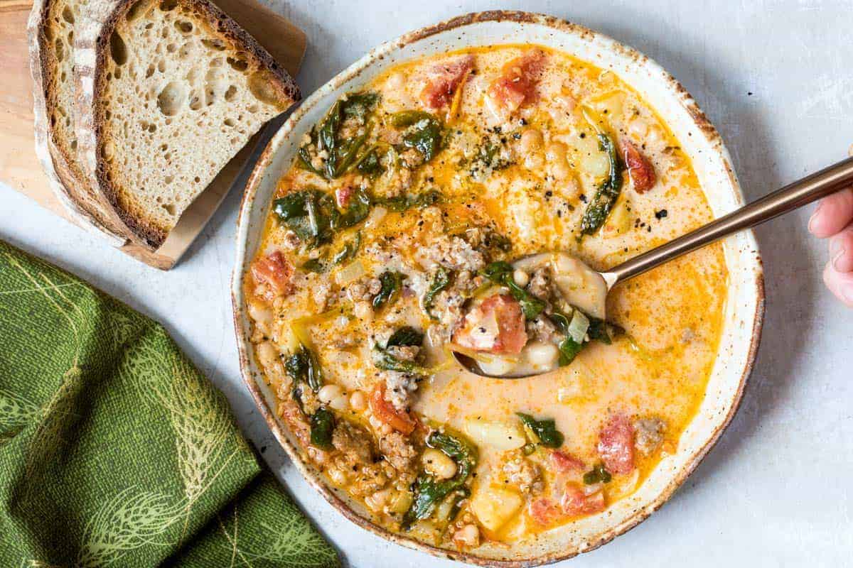 bowl of tuscan bean soup with spoon in it next to slices of bread and napkin