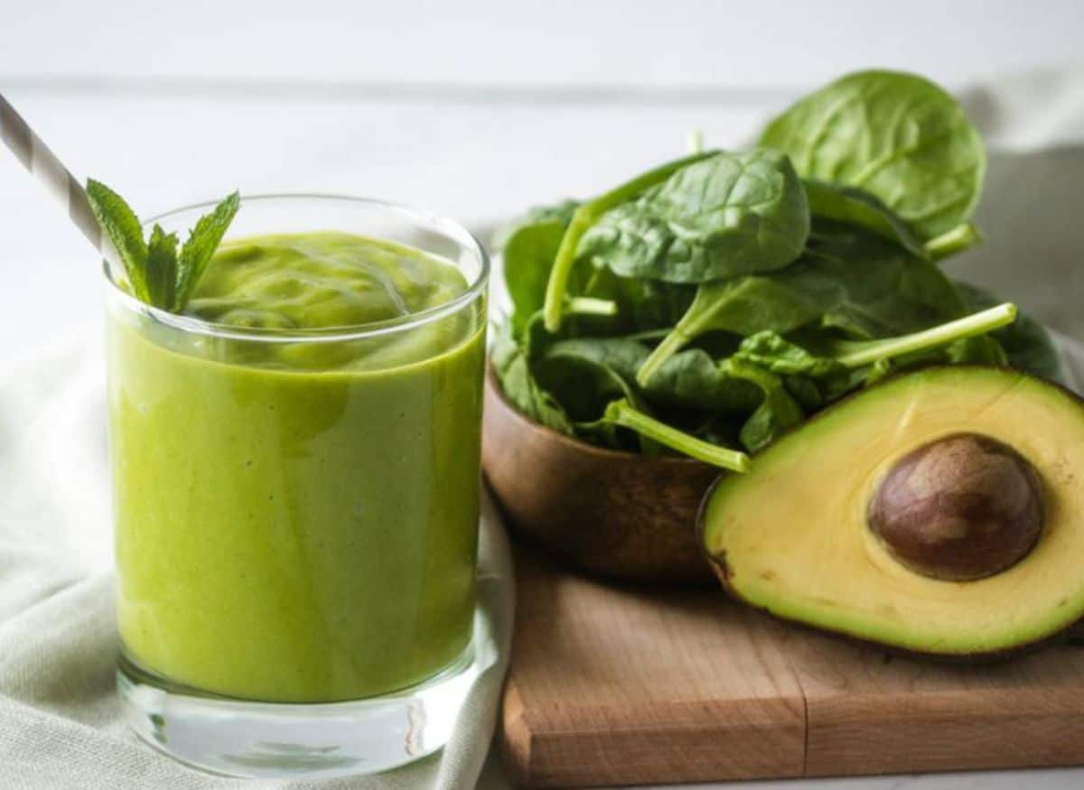 Green smoothie in a glass with spinach and half an avocado in the background.