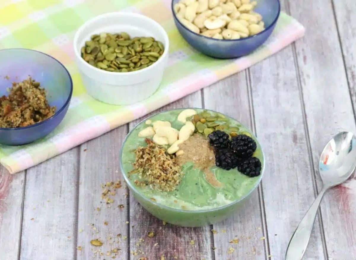 Green avocado smoothie bowl topped with nuts and berries with a spoon beside it and toppings behind it.