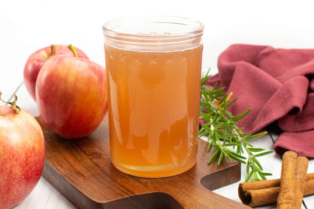 A glass of apple cider with cinnamon sticks and apples on a cutting board.