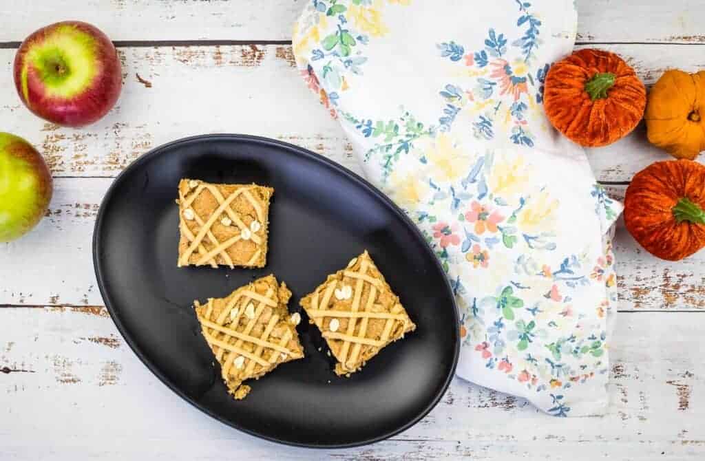 A black plate with Brown Butter Apple Blondie squares with apples on the left and pumpkins on the right side of the table.