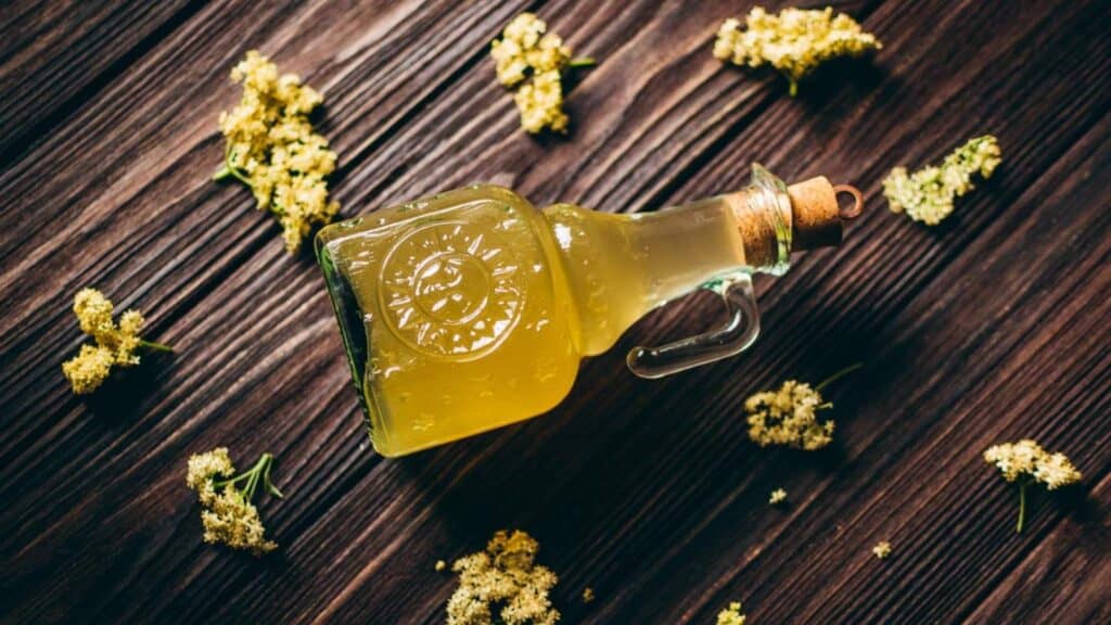 A bottle of elderflower cordial surrounded by flowers and edible gifts on a wooden table.