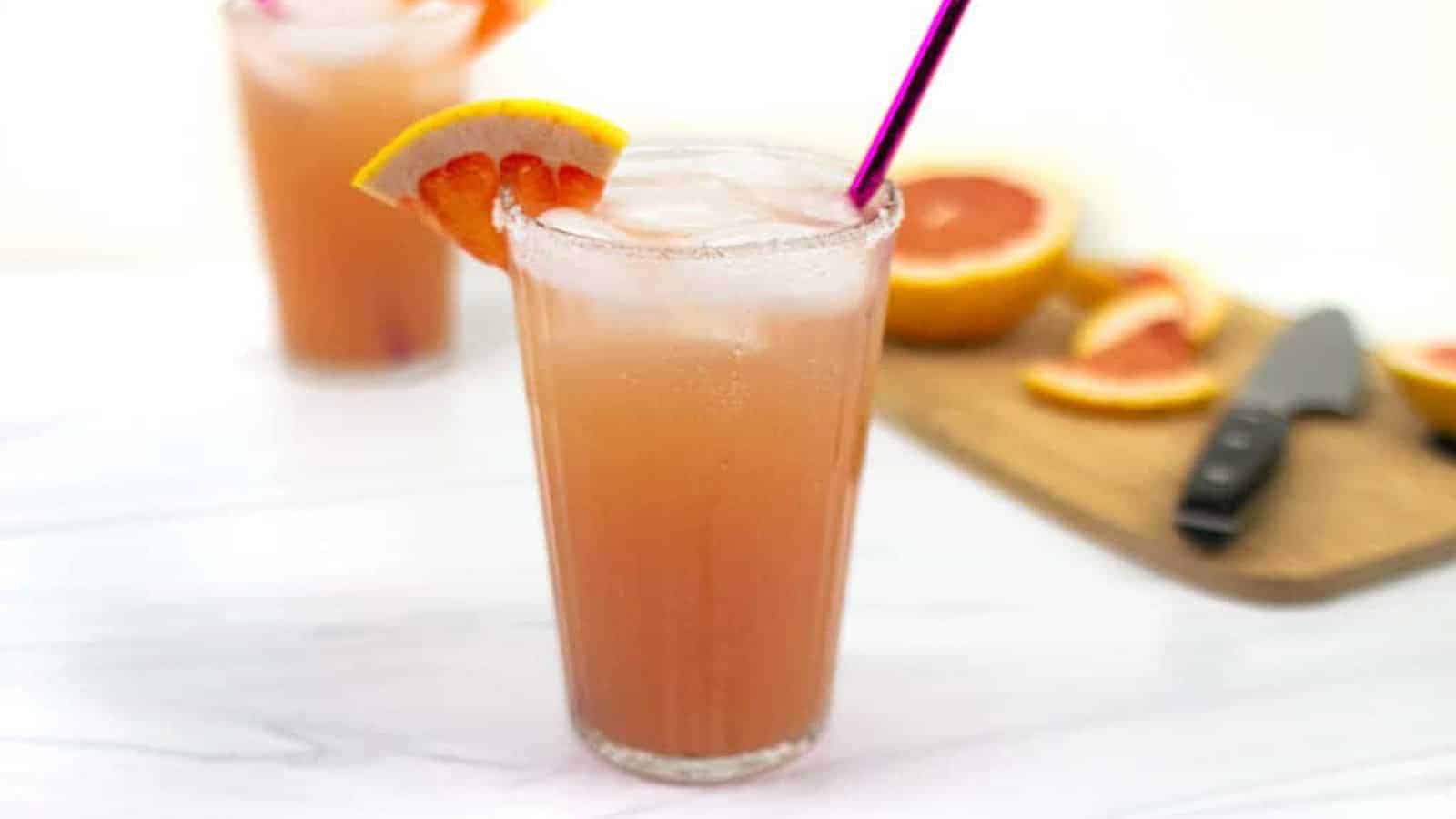 Two pink cocktails garnished with a wedge of grapefruit sit alongside a cut grapefruit on a cutting board with a knife.