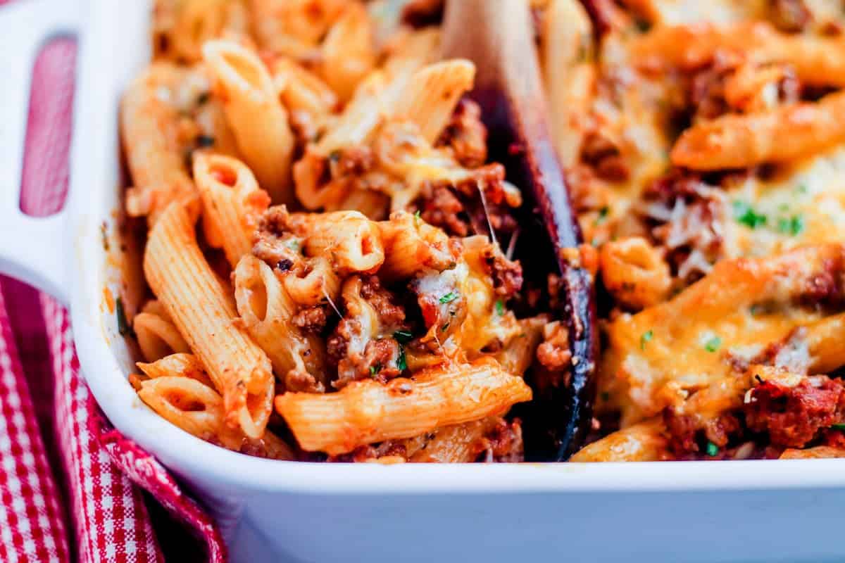 A spoon scooping a portion of penne pasta bake from a casserole dish.