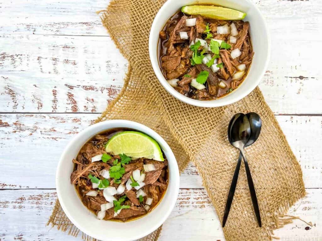 Two white bowls filled with slow cooker Birria garnished with fresh lime wedges.