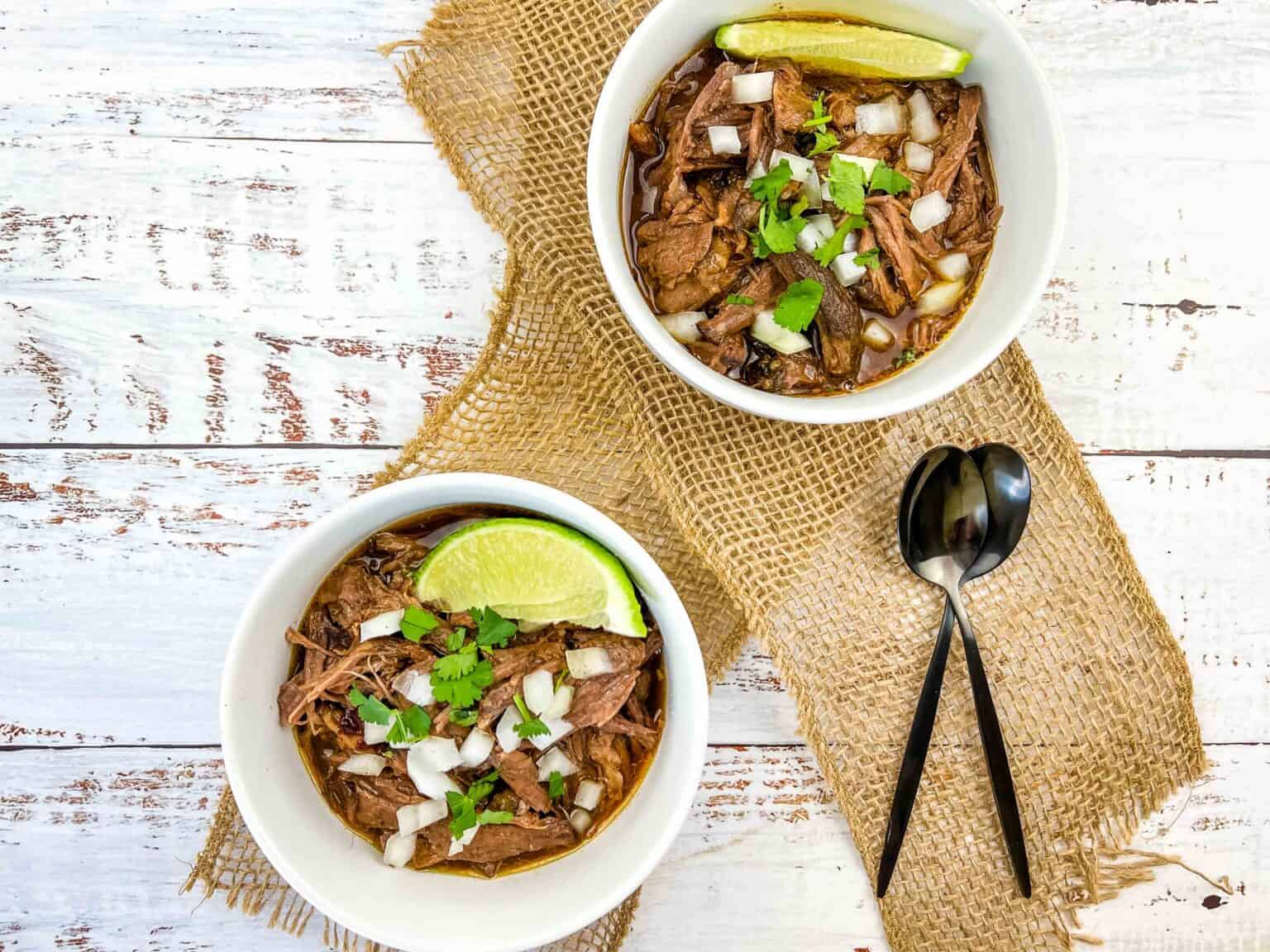 Two white bowls filled with slow cooker birria garnished with fresh lime wedges.