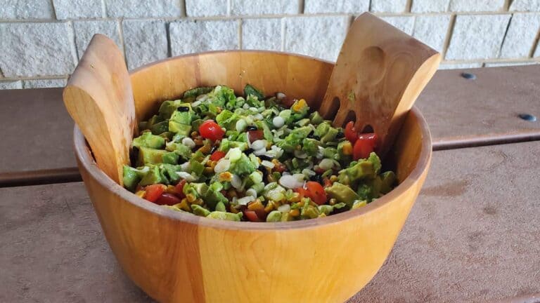 Image shows a wooden bowl containing avocado tomato salad shown from overhead and sitting on a picnic table.