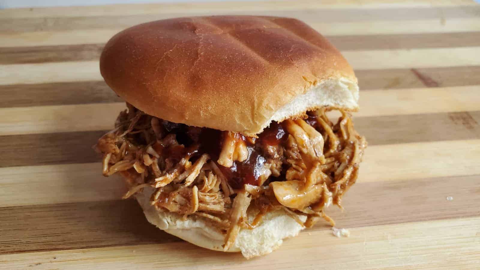 Image shows a Pulled pork sandwich on white plate with pretzels on the side.