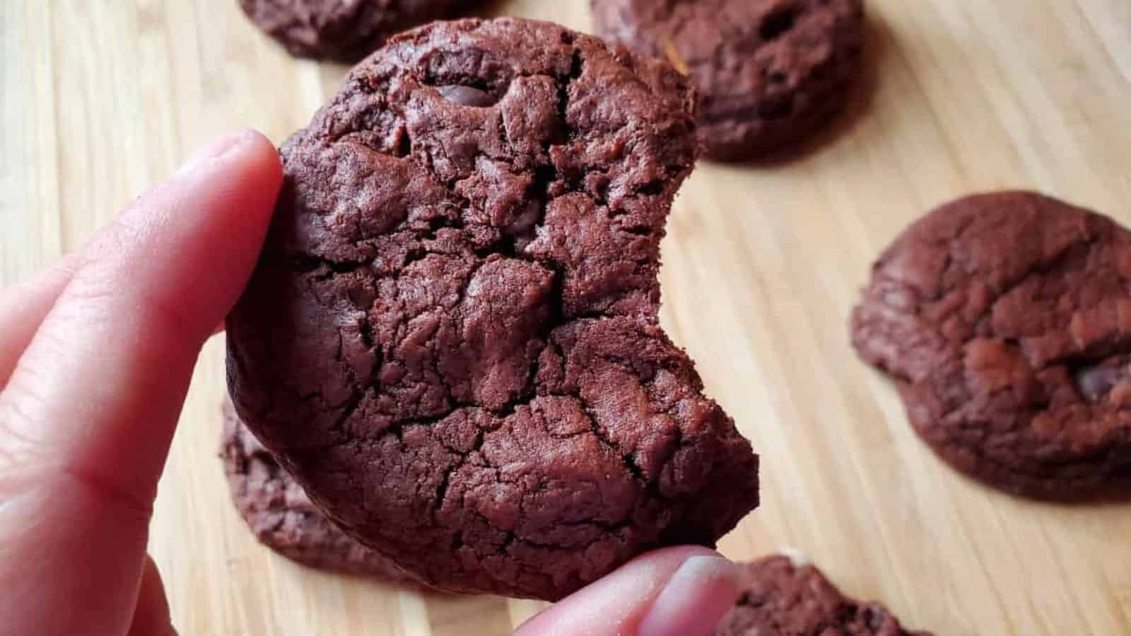 Image shows shows a hand holding up a chocolate cookie with a bite taken out of it and more in the background.