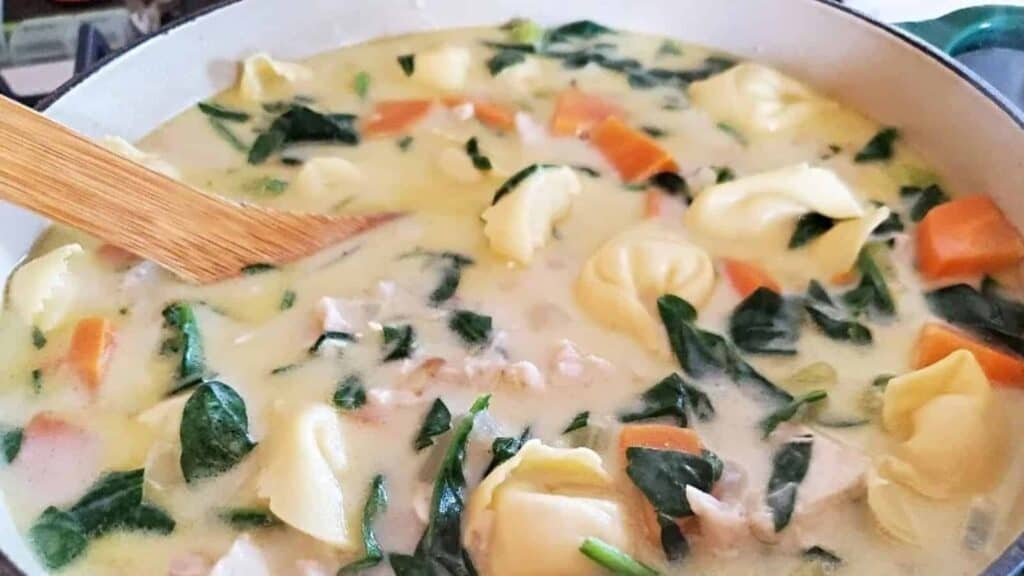 Image shows Chicken tortellini soup in a ceramic enameled pot with a wooden spoon stirring it.