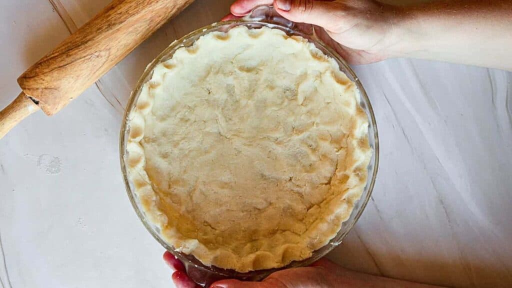 A person rolling out dough for a pie crust in a glass dish.