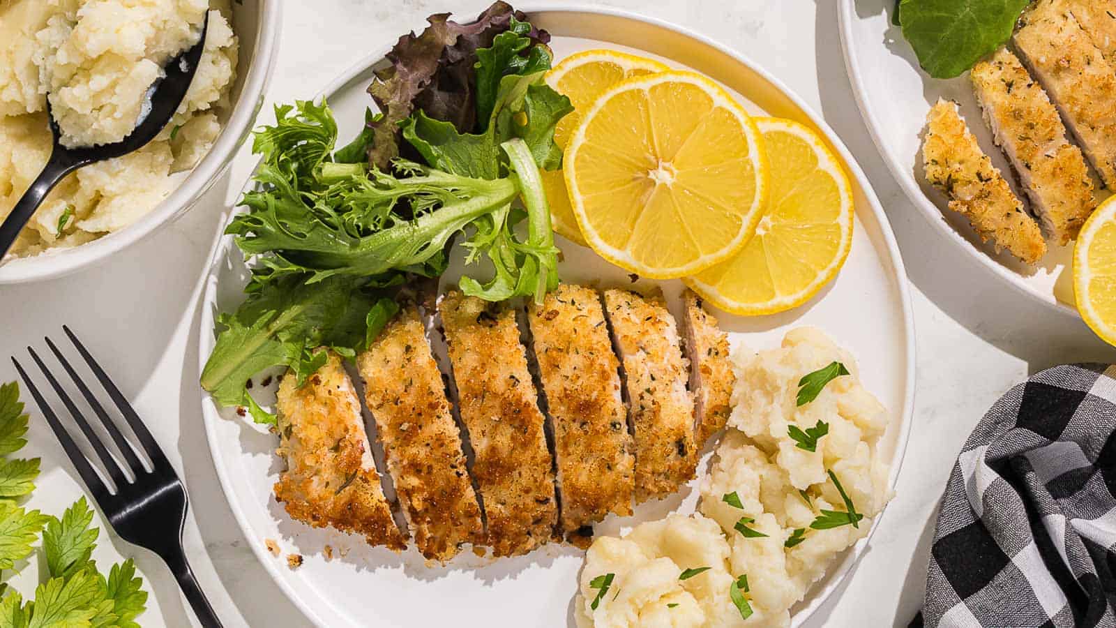 Breaded italian chicken cutlets on a black plate with sliced lemon.