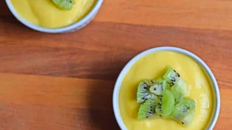 An overhead shot shows two ramekins with mango custard topped with chopped kiwi on a wooden cutting board.