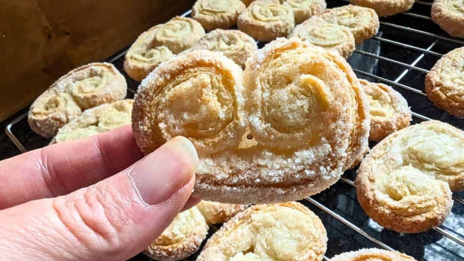 Image shows a hand holding a palmier cookie in front of others on a wire cooling rack.