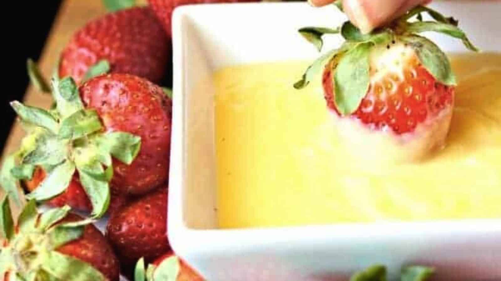 Image shows  A person dipping strawberries into a bowl of pineapple curd.