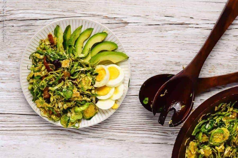 A serving of shaved brussels sprouts salad next to the serving bowl of shaved brussels sprouts salad with salad servers in the middle.