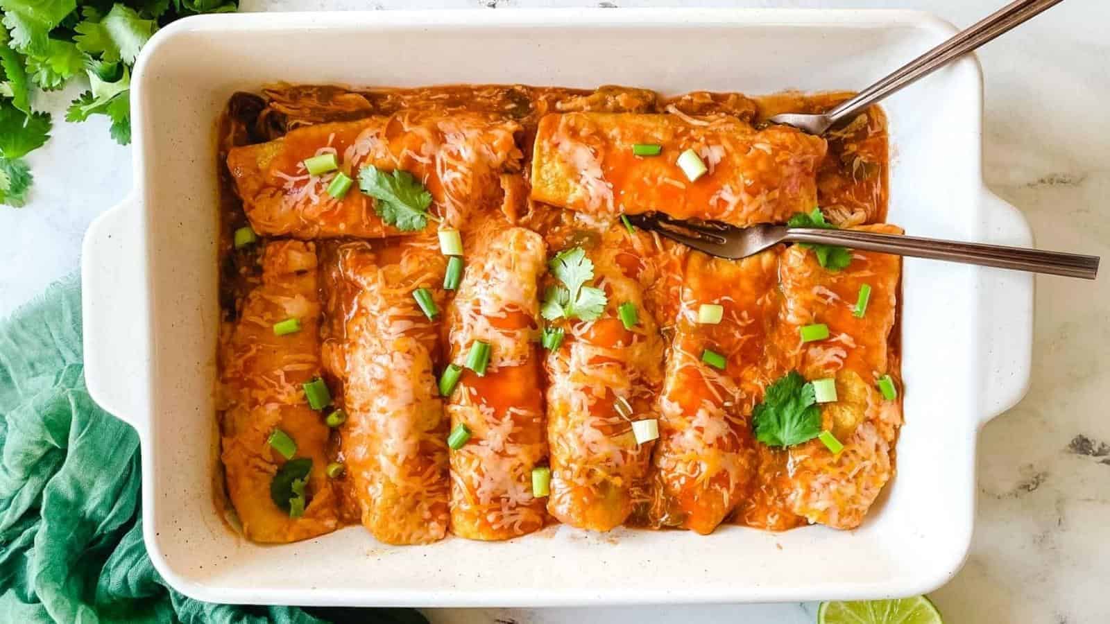 A baking dish full of chicken enchiladas is surrounded by cut limes, a halved avocado, and a green linen.