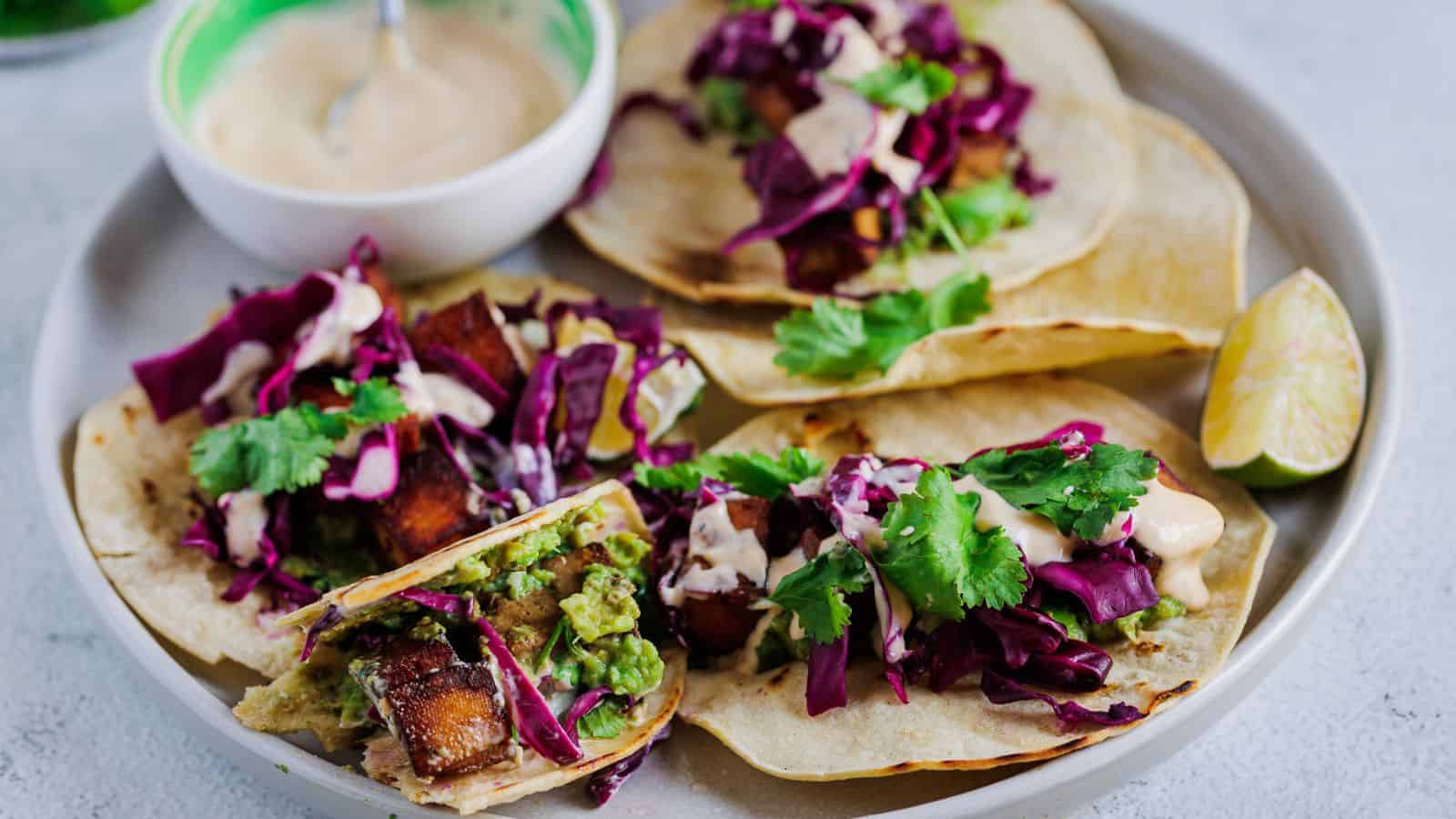 Five tacos with tofu and dipping sauce on a plate.