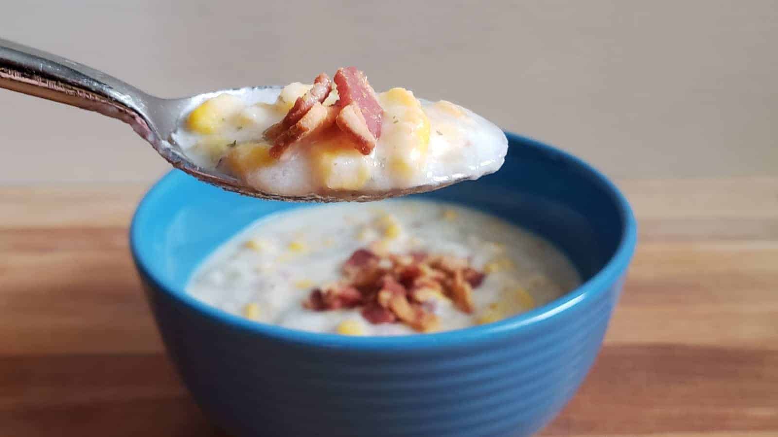 Spoon holding potato and corn chowder with bacon with the full bowl of soup behind it.