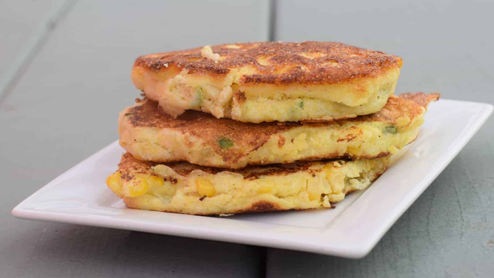 Image shows three corn fritters stacked atop each other on a white plate with a blue wooden background.