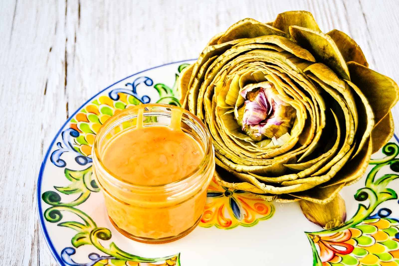 A holiday appetizer, an artichoke on a plate, is elegantly served next to a jar of sauce.